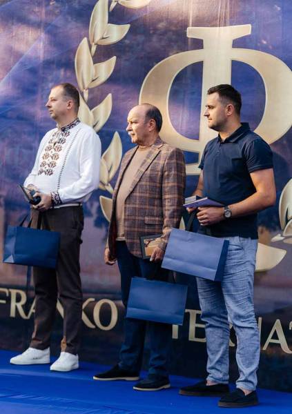 Троє чоловіків із нагородами та синіми пакунками на сцені під час вручення Міжнародної премії імені Івана Франка у Дрогобичі.
